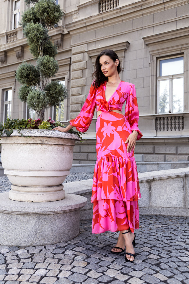 Frau in einem pink-roten Maxikleid mit floralem Muster, Cut-out-Detail und Volantrock vor historischer Kulisse.