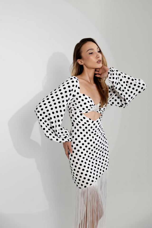 Frau in einem gepunkteten, figurbetonten Kleid mit Cut-outs, langen Puffärmeln und Fransen, elegant posierend.