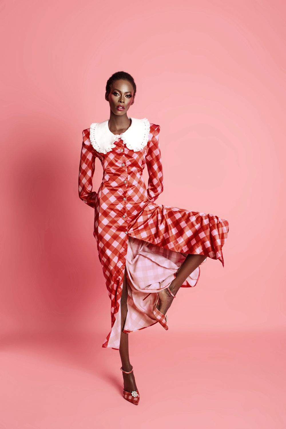 Langes kariertes Kleid mit Knopfleiste, weißen Manschetten und Kragen, getragen von einem Model vor rosa Hintergrund.
