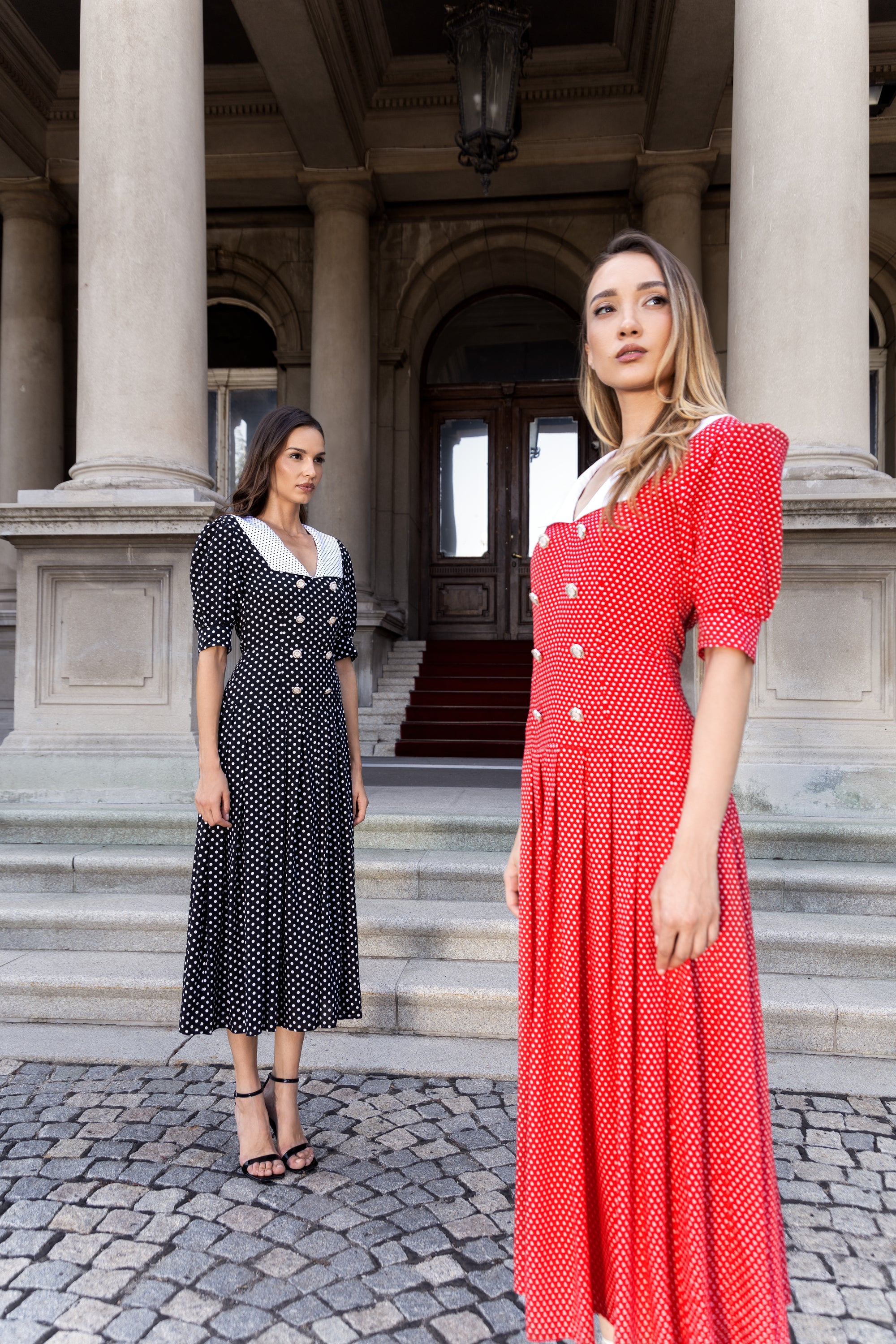 Zwei Frauen in gepunkteten Midikleidern mit weißem Kragen, Doppelreiher-Knöpfen und Puffärmeln vor historischer Kulisse.